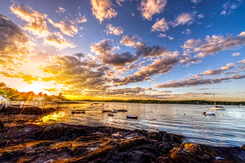 Sunset over a calm bay with boats, rocky shore, and vibrant clouds reflecting warm colors in the sky.