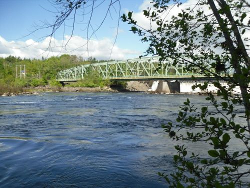 A green bridge spans a river, surrounded by trees and a clear blue sky with scattered clouds.