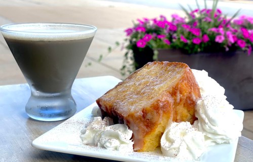 A slice of glazed cake topped with whipped cream, served alongside a glass of coffee, with flowers in the background.