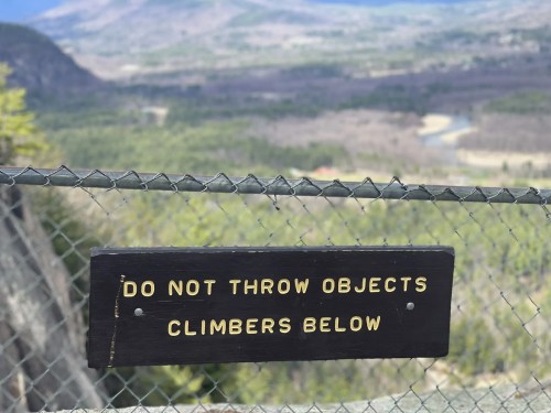 Sign on a fence warning against throwing objects, with a scenic view of a valley and mountains in the background.