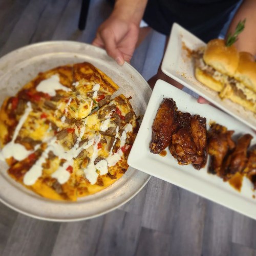 A person holds a large pizza topped with meat and drizzled sauce, alongside a plate of chicken wings and a sandwich.