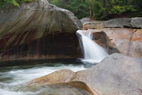 A serene waterfall cascades over smooth rocks, surrounded by lush greenery in a tranquil forest setting.