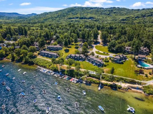 Aerial view of a lakeside resort with boats, green trees, and hills in the background under a clear blue sky.