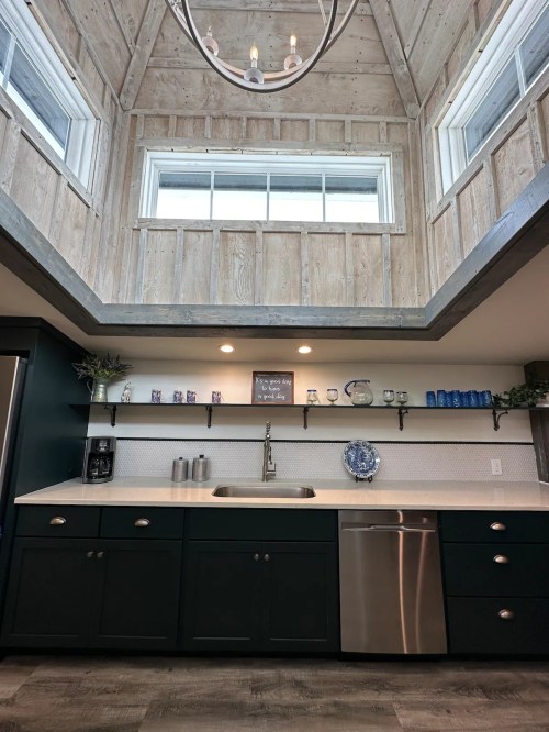 Modern kitchen with dark cabinets, a stainless steel sink, and a high wooden ceiling with large windows.