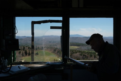 A person reading a map inside a lookout tower, with a scenic view of mountains and forests through the window.