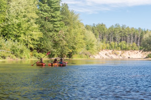 Two people kayaking on a calm river surrounded by lush green trees and a clear blue sky.