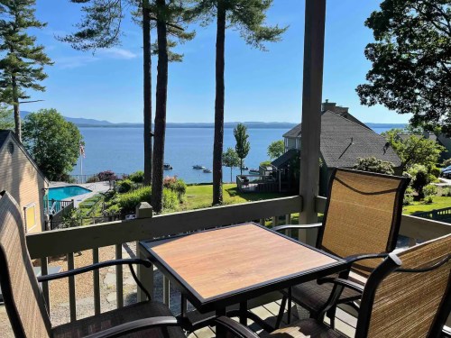 A serene lakeside view from a porch, featuring a table and chairs, surrounded by trees and houses.