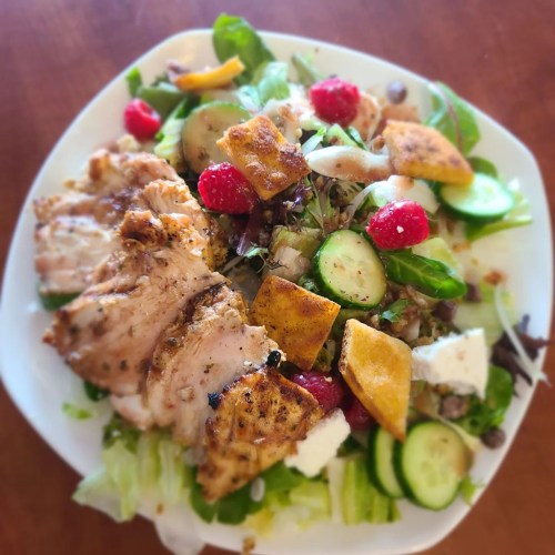 A colorful salad topped with grilled chicken, cucumbers, raspberries, and croutons on a white plate.