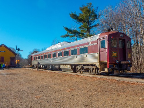A vintage train car parked at a station surrounded by trees under a clear blue sky.