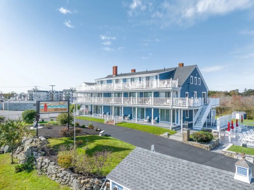 A blue multi-story building with balconies, surrounded by green grass and a clear sky, near a coastal area.