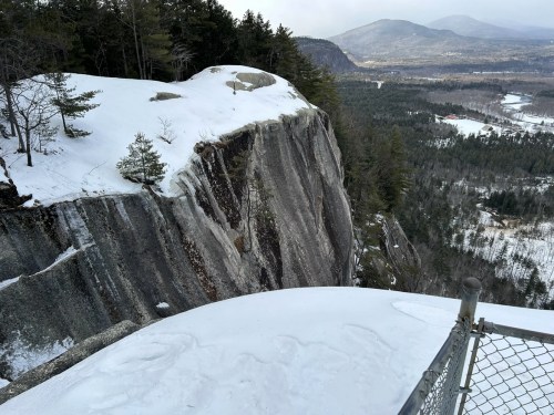 Snow-covered cliff edge with trees, overlooking a valley and mountains in the distance.