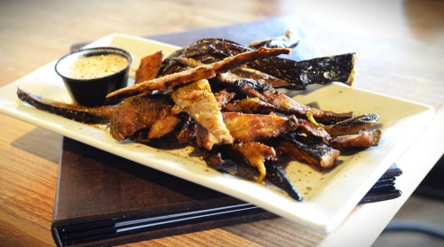A plate of crispy fried fish strips served with a small bowl of dipping sauce on a wooden table.