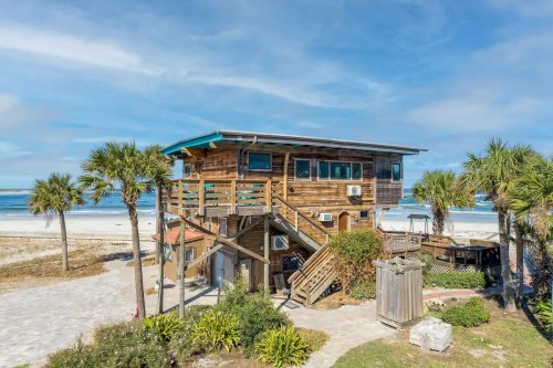 A wooden beach house with stairs, surrounded by palm trees, overlooking a sandy beach and ocean under a blue sky.