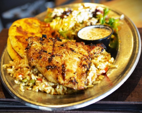 A plate of grilled chicken with rice, a side salad, and a small cup of sauce, served with flatbread.