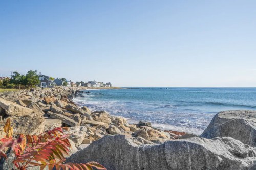 A serene coastal view with rocky shoreline, calm blue water, and distant houses under a clear sky.