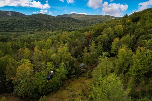 A person zip-lining over a lush, green forest with mountains in the background under a partly cloudy sky.