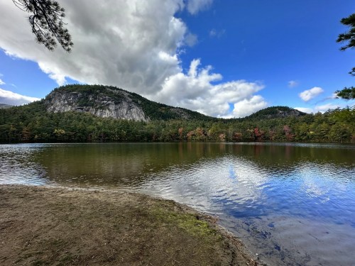 A serene lake surrounded by mountains and trees, with a blue sky and fluffy clouds reflecting on the water's surface.