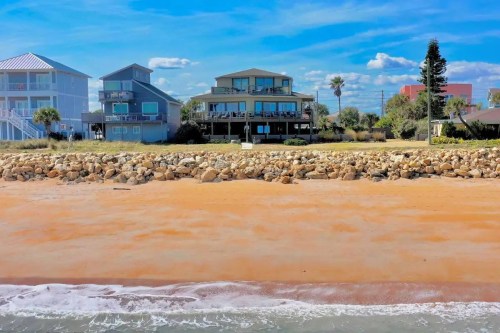 A sandy beach with rocky shoreline and colorful houses in the background under a bright blue sky.