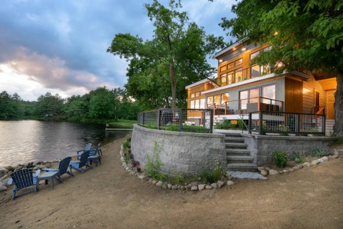A modern lakeside home with large windows, surrounded by trees, and a sandy area with blue chairs by the water.