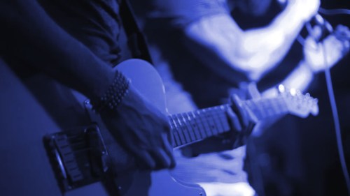 Close-up of a guitarist's hands playing an electric guitar, illuminated in blue light.