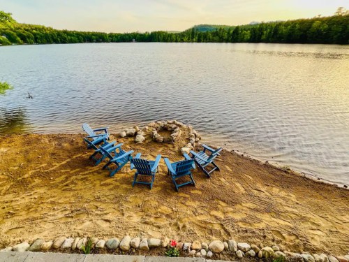 A serene lakeside scene with blue chairs arranged around a stone fire pit on a sandy shore.