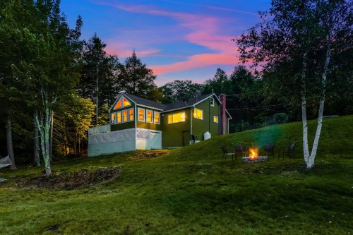 A cozy green house at dusk, surrounded by trees, with a fire pit in the foreground and colorful sky above.