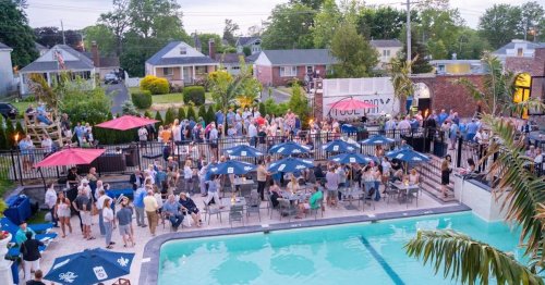 A lively outdoor gathering by a pool, with people socializing under umbrellas and a backdrop of houses and greenery.