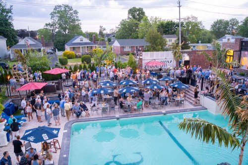 A lively outdoor gathering by a pool, with people socializing under umbrellas and a backdrop of homes and trees.