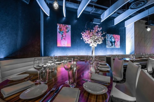 Elegant restaurant interior with a long table set for dining, featuring artistic wall decor and a floral centerpiece.