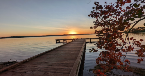 A wooden dock extends into a calm lake at sunset, with colorful leaves framing the scene.