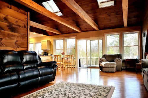 Cozy living room with wooden beams, a black leather couch, and large windows overlooking a green outdoor view.