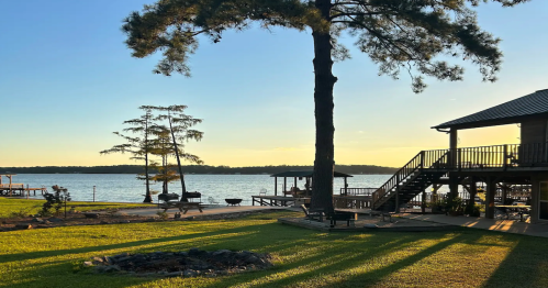 A serene lakeside view at sunset, featuring trees, a wooden deck, and a fire pit on the grassy shore.