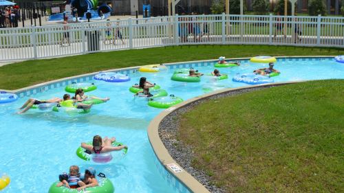 A lazy river with children floating on colorful inner tubes, surrounded by green grass and a white fence.