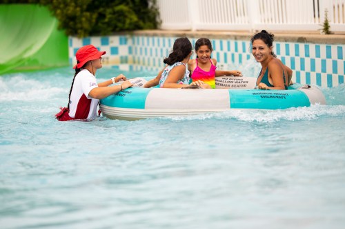 A lifeguard assists a family on a float in a water park, with children enjoying the ride in a pool setting.