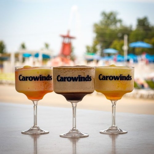Three colorful cocktails in large glasses with "Carowinds" logo, set against a sunny outdoor background.