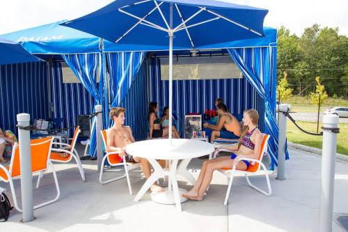A group of young people relaxes in cabanas by a pool, sitting at a table under blue umbrellas.