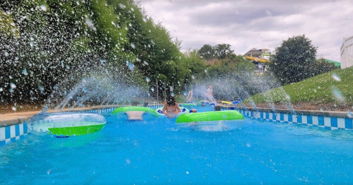 A splash-filled pool scene with colorful floats and children playing, surrounded by greenery and a cloudy sky.