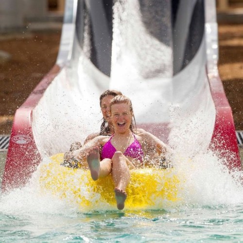 Two women slide down a water slide on a yellow inner tube, splashing into a pool below.