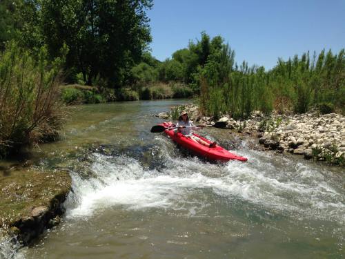 A person in a red kayak navigates a rocky river with greenery on the banks under a clear blue sky.