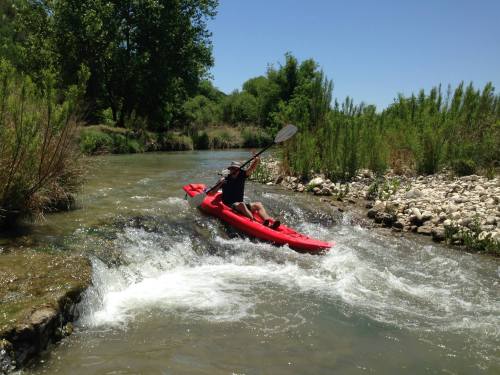 A person kayaking on a river, navigating through small rapids surrounded by greenery and rocks.