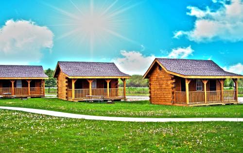 Three wooden cabins on a grassy area, with a bright blue sky and sun shining above.