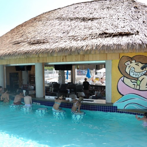 A swim-up bar with patrons enjoying drinks in a pool, featuring a colorful mural of a smiling lion.