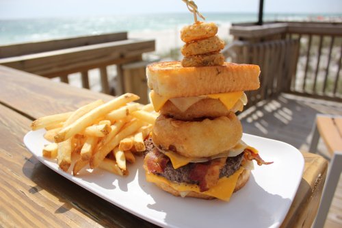 A towering burger with cheese, bacon, and a donut, served with crispy fries on a beachside table.