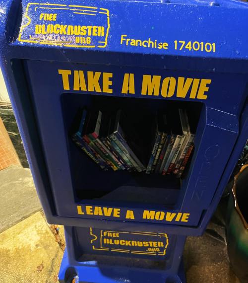 A blue Blockbuster movie rental box with the text "Take a movie, Leave a movie" and shelves of DVDs inside.
