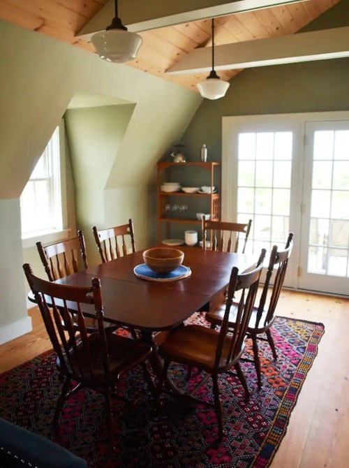 A cozy dining area with a wooden table, six chairs, and a colorful rug, featuring large windows and a shelf of dishes.