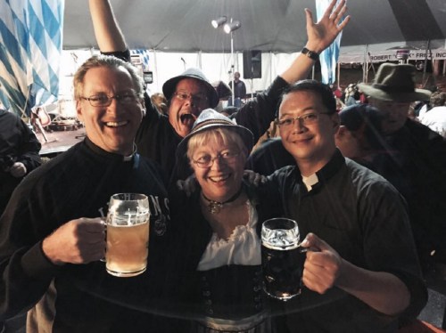 Four people in festive attire hold large beer mugs, smiling and celebrating at a lively outdoor event.