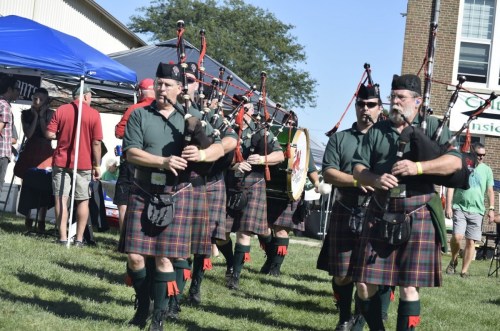 A group of men in kilts playing bagpipes at an outdoor event, with tents and spectators in the background.