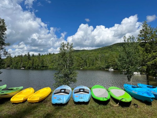 Colorful kayaks lined up by a serene lake, surrounded by lush green trees and a blue sky with fluffy clouds.