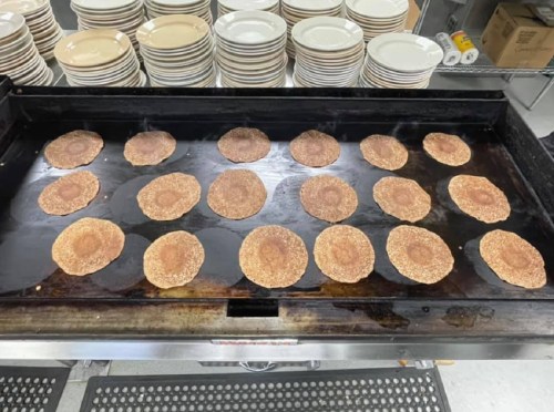 A griddle with multiple pancakes cooking, surrounded by stacks of plates in a kitchen setting.