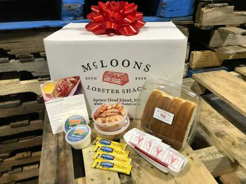 A gift box from McLoons Lobster Shack containing lobster, rolls, butter, and condiments, wrapped with a red bow.